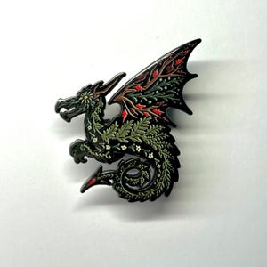 May include: A black enamel pin featuring a green dragon with floral details. The dragon's wings are spread and have a red and green floral pattern. The dragon's body is covered in green leaves and white flowers.