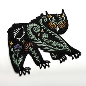 May include: Black embroidered patch featuring a stylized bear-owl hybrid design. The patch has a detailed floral and leaf pattern in green, white, and orange. The owl's eyes are yellow. The patch is on a white background.