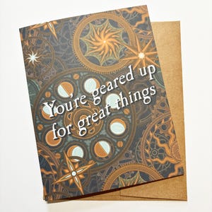 May include: A card with a brown, gold, and white patterned design featuring gears and celestial bodies. The text "You're geared up for great things" is printed in white on the card.
