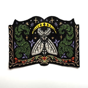 Puede incluir: Parche negro bordado con un diseño de polilla con alas blancas detalladas, sobre un fondo de estrellas, luna creciente y elementos florales. El diseño incluye follaje verde, flores moradas y detalles dorados y naranjas, creando una estética mística.