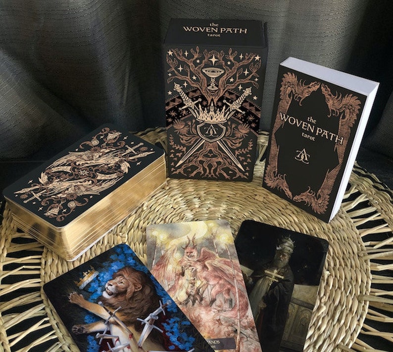 May include: A tarot deck box with gold accents and the text "The Woven Path Tarot" on the front. The box is open and shows the cards inside. The cards have illustrations of a lion, a winged figure, and a person in a robe.