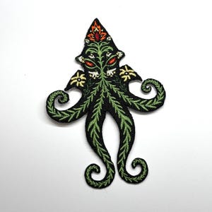 Puede incluir: Parche bordado con forma de pulpo con detalles florales y de hojas. El parche presenta un fondo negro con detalles en verde, amarillo, blanco y rojo. Los tentáculos tienen un diseño rizado.