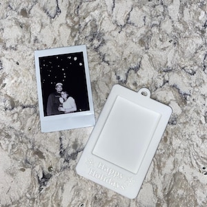 Peut inclure: Une photo Polaroid en noir et blanc d'un couple, à côté d'un cadre photo blanc avec l'inscription "Happy Holidays" et des motifs de flocons de neige. Le cadre est percé d'un trou pour l'accrochage.