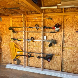 May include: A wooden wall-mounted storage rack with multiple shelves holding various gardening tools, including weed trimmers, a rake, and a long-handled tool. The rack is installed in a wooden shed.