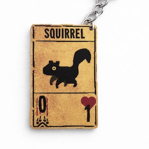 May include: A keychain with a rectangular tag displaying a black squirrel illustration and the word "SQUIRREL". The tag has a yellow background with a heart and the number "1". The keychain has a silver metal ring.