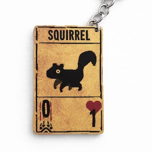 Puede incluir: Un llavero rectangular con un diseño de tarjeta color canela y una ilustración de ardilla negra pixelada. El texto SQUIRREL aparece en la parte superior, con un cero y una huella en la parte inferior izquierda, y un corazón rojo con el número uno.