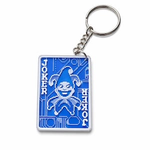 Puede incluir: Un llavero rectangular con un diseño de arte en píxeles azul y blanco de un personaje de bufón. La palabra JOKER está impresa verticalmente a ambos lados de la figura central. El llavero incluye una anilla de metal plateado con una cadena corta.