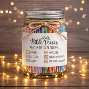 May include: A glass jar with a metal lid, filled with coloured paper strips and tied with twine. The label reads "Bible Verses" and lists feelings. The background has fairy lights. The jar is filled with colourful paper strips.