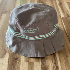 May include: A brown bucket hat with a white and light green striped band. The hat has the text "adidas SAFETY" in white with a black adidas logo.