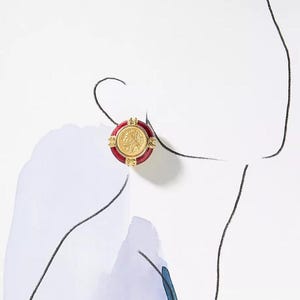 May include: A gold-colored coin earring with a red border. The coin features a profile image and is surrounded by a decorative gold frame. The earring is round and has a post backing.