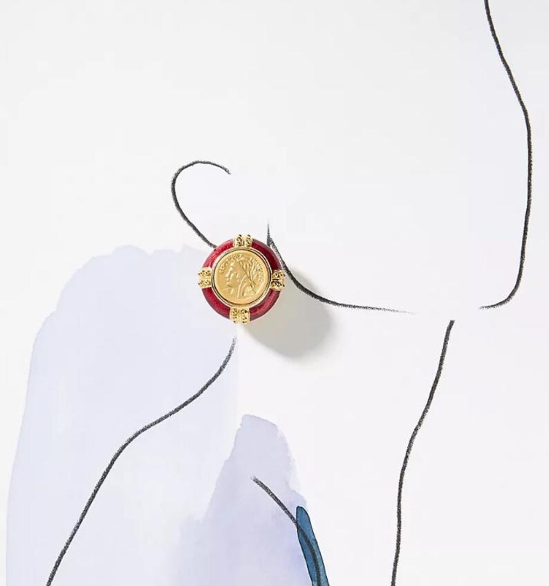May include: A gold-colored coin earring with a red border. The coin features a profile image and is surrounded by a decorative gold frame. The earring is round and has a post backing.
