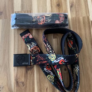 May include: A black belt with a colorful tattoo-inspired design featuring roses, hearts, and a woman's face. The belt has a black buckle with the Ed Hardy logo.