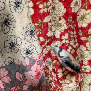 May include: A close-up of a patchwork textile featuring floral patterns. The design incorporates sections of red, cream, and gray fabrics, each adorned with different floral motifs in shades of white, pink, and navy blue. The textile has a soft, textured appearance.