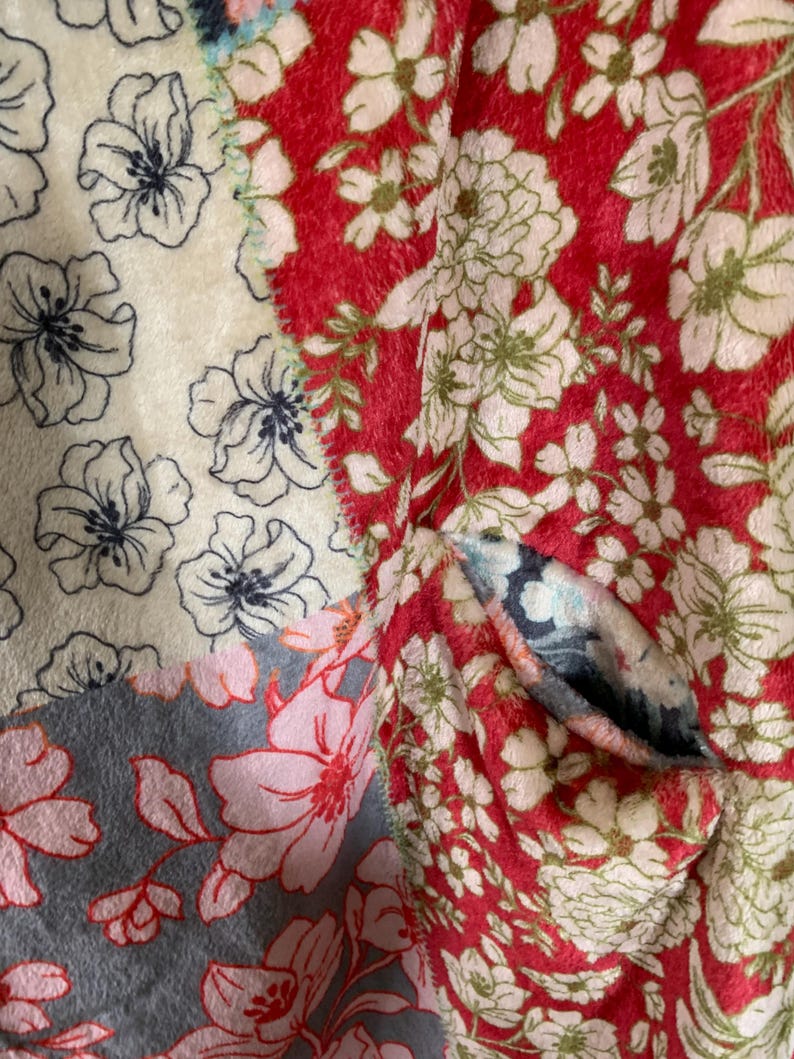May include: A close-up of a patchwork textile featuring floral patterns. The design incorporates sections of red, cream, and gray fabrics, each adorned with different floral motifs in shades of white, pink, and navy blue. The textile has a soft, textured appearance.
