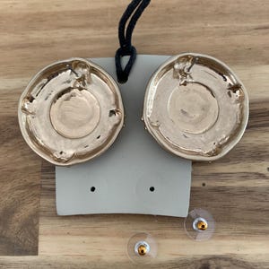 May include: Gold-toned, round earrings with a textured design. The earrings are displayed on a gray card with two clear earring backs. A black cord is attached to the card. The earrings are on a wooden surface.
