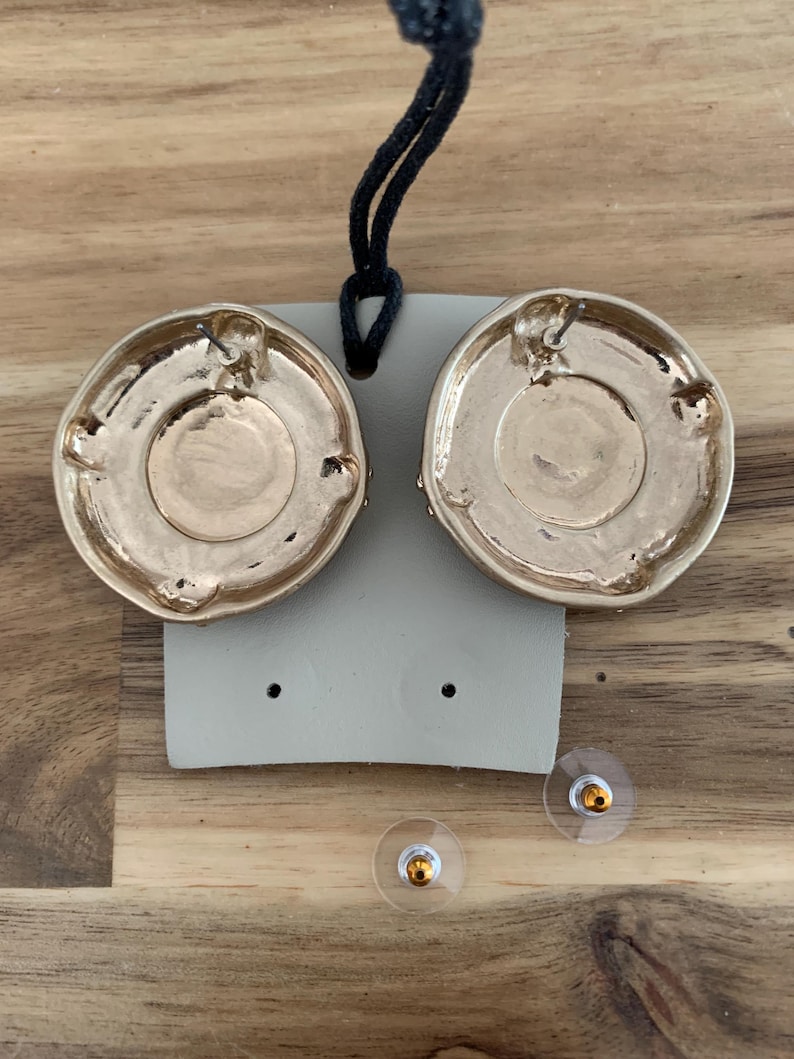 May include: Gold-toned, round earrings with a textured design. The earrings are displayed on a gray card with two clear earring backs. A black cord is attached to the card. The earrings are on a wooden surface.