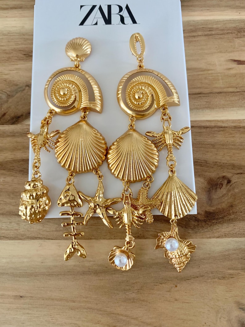 Puede incluir: Pendientes dorados con colgantes de conchas marinas, estrellas de mar y langostas. Los pendientes tienen un dise&ntilde;o en espiral en la parte superior y un acento de perla. El logotipo de ZARA es visible en el fondo.