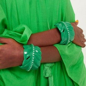 May include: Two green leaf-shaped bracelets with white dot accents. The bracelets are worn on both wrists. The person is wearing a green outfit. The bracelets have a glossy finish and are a vibrant shade of green.