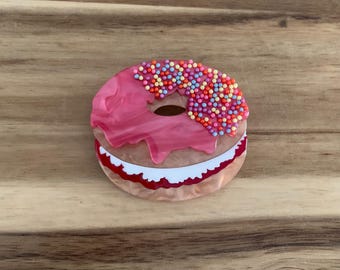 Puede incluir: Broche en forma de donut rosa con acabado brillante. La capa superior presenta glaseado rosa con chispas de colores. La capa inferior tiene un diseño blanco y rojo. El broche es un accesorio divertido.