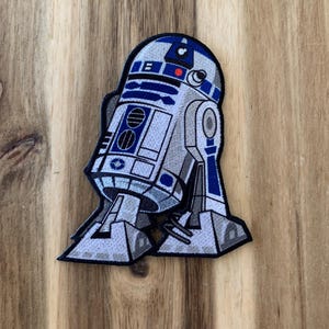 May include: Embroidered patch featuring the iconic R2-D2 droid from Star Wars. The patch showcases the droid's signature blue, white, and gray color scheme, with detailed stitching. The patch is a collectible item for fans of the Star Wars franchise.