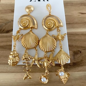 Puede incluir: Pendientes dorados con tem&aacute;tica de conchas marinas. Cada pendiente presenta una gran concha en espiral, una concha de vieira y varios abalorios, incluyendo una estrella de mar, un esqueleto de pez y una perla. Los pendientes son de la marca ZARA.