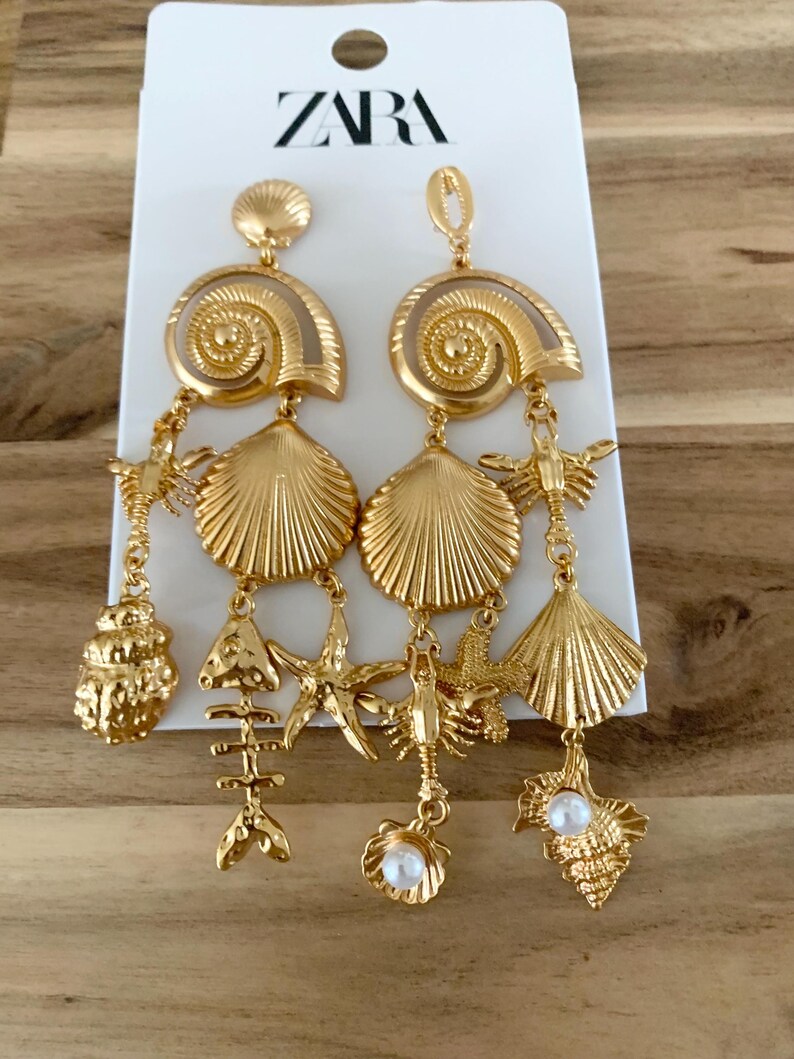 Puede incluir: Pendientes dorados con tem&aacute;tica de conchas marinas. Cada pendiente presenta una gran concha en espiral, una concha de vieira y varios abalorios, incluyendo una estrella de mar, un esqueleto de pez y una perla. Los pendientes son de la marca ZARA.