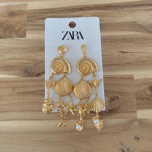 Puede incluir: Pendientes dorados con colgantes de conchas marinas y vida marina. Los pendientes tienen una secci&oacute;n superior con un dise&ntilde;o en espiral, seguida de varios colgantes, incluyendo conchas, estrellas de mar y un esqueleto de pez. Los pendientes est&aacute;n sujetos a una etiqueta blanca con la marca "ZARA".