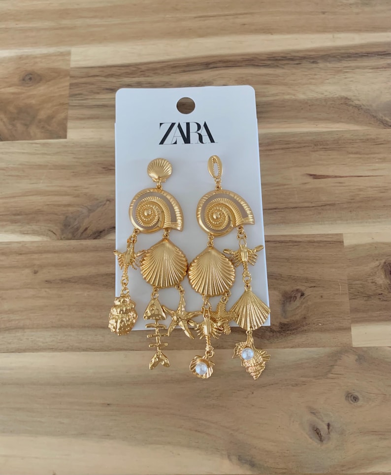 Puede incluir: Pendientes dorados con colgantes de conchas marinas y vida marina. Los pendientes tienen una secci&oacute;n superior con un dise&ntilde;o en espiral, seguida de varios colgantes, incluyendo conchas, estrellas de mar y un esqueleto de pez. Los pendientes est&aacute;n sujetos a una etiqueta blanca con la marca "ZARA".