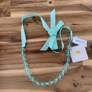 Puede incluir: Un collar de cuentas azul claro con una cinta azul claro a juego atada en un lazo. El collar está sobre una superficie de madera y tiene una etiqueta que dice "Crew Cuts Factory Store".