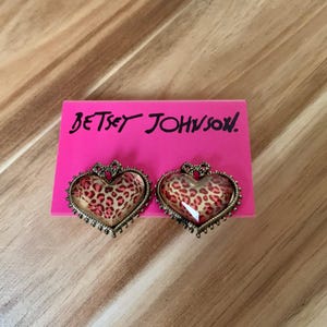 Könnte beinhalten: Ein Paar goldene herzförmige Ohrringe mit Leopardenmuster. Die Ohrringe befinden sich auf einer rosa Karte mit dem Text "Betsey Johnson!" in Weiß.