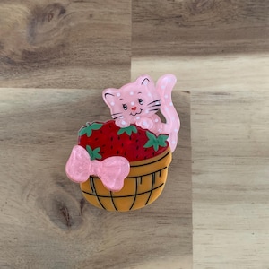 May include: A pink cat with white polka dots peeks over a basket of strawberries. The basket is yellow with a pink bow and green leaves. The strawberries are red.