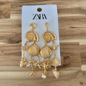 Puede incluir: Pendientes dorados con tem&aacute;tica de conchas marinas. Los pendientes tienen un dise&ntilde;o en espiral en la parte superior, con colgantes que incluyen conchas, estrellas de mar y espinas de pescado. La etiqueta de la marca dice "ZARA".