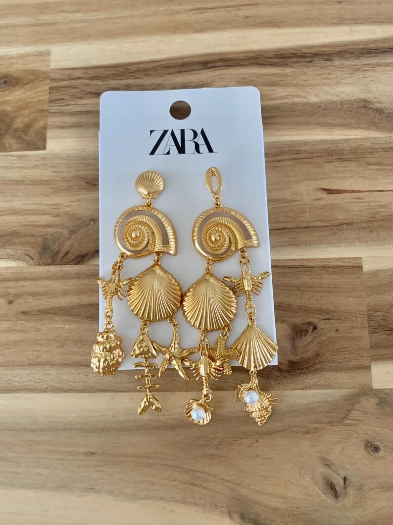 Puede incluir: Pendientes dorados con tem&aacute;tica de conchas marinas. Los pendientes tienen un dise&ntilde;o en espiral en la parte superior, con colgantes que incluyen conchas, estrellas de mar y espinas de pescado. La etiqueta de la marca dice "ZARA".