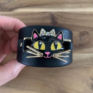 Può includere: Bracciale a polsino in pelle nera con un design a forma di gatto nero. Il gatto ha occhi gialli, naso rosa e baffi dorati, e un fiocco gioiello. Il bracciale ha una fibbia dorata e una chiusura a scatto. Circa 5 cm di larghezza.