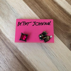 May include: A pair of Betsey Johnson earrings. One earring features a red square-shaped gem with a gold-tone setting. The other earring features a gold-tone rabbit with a pearl accent.