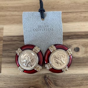 May include: A pair of red and gold-toned clip-on earrings. Each earring features a gold coin design with a profile of a person, surrounded by a red border and embellished with small, clear crystals. A gray tag with "URBAN OUTFITTERS" is attached.