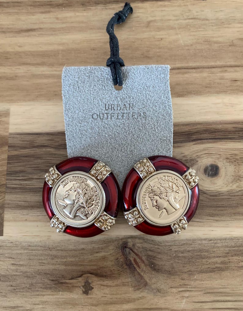 May include: A pair of red and gold-toned clip-on earrings. Each earring features a gold coin design with a profile of a person, surrounded by a red border and embellished with small, clear crystals. A gray tag with "URBAN OUTFITTERS" is attached.