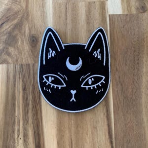 Könnte beinhalten: Schwarzer Katzenaufnäher mit weißen Akzenten. Das Gesicht der Katze hat einen Mond über den Augen und eine kleine Nase. Ein einzigartiges Accessoire für Kleidung oder Taschen.