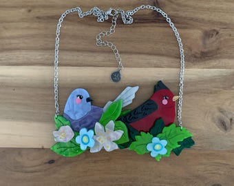 Puede incluir: Collar de cadena plateada con un colgante que representa dos pájaros coloridos sobre hojas y flores verdes. Un pájaro es azul, el otro es rojo. El collar tiene una cadena ajustable.