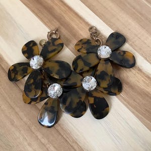 May include: A pair of flower-shaped earrings with a tortoiseshell pattern in shades of brown and black. Each earring features a large, clear, round crystal in the center and a smaller crystal below. The earrings are set against a wooden background.