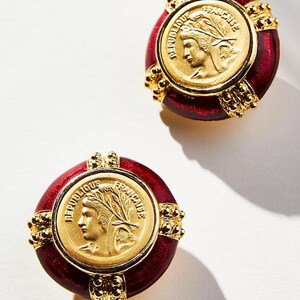May include: A pair of gold-toned earrings with a red enamel border. Each earring features a gold coin with a profile and the words "REPUBLIQUE FRANCAISE". The earrings have a vintage aesthetic.