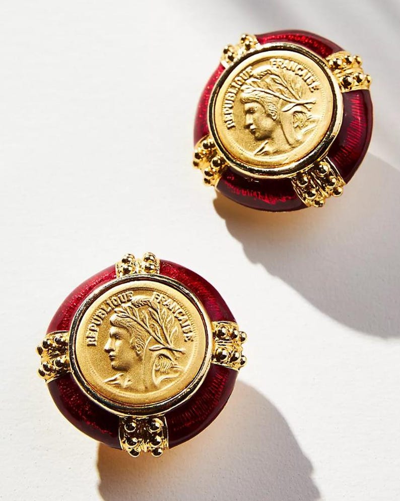 May include: A pair of gold-toned earrings with a red enamel border. Each earring features a gold coin with a profile and the words "REPUBLIQUE FRANCAISE". The earrings have a vintage aesthetic.