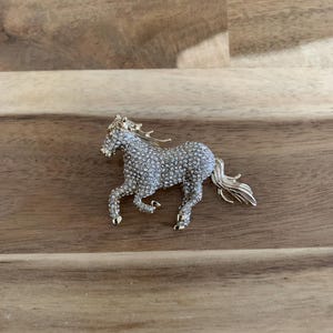 May include: A gold-toned horse brooch with a clear crystal design. The horse is running and has a flowing mane and tail.