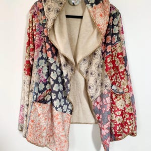 May include: A patchwork floral print jacket with a beige interior lining. The jacket features a shawl collar, two front pockets, and a stitched trim. The floral patterns include red, pink, blue, and beige colors. The jacket is hanging on a black hanger.