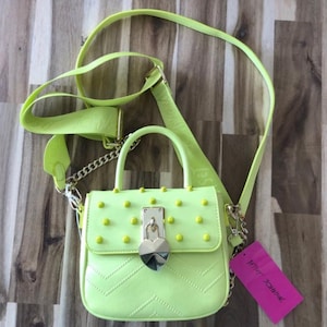 May include: A neon yellow handbag featuring a top handle, a detachable shoulder strap, and a front flap adorned with small matching studs. The bag includes a silver-toned heart-shaped padlock closure and a pink brand tag attached to the side.