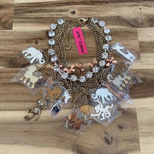 May include: A gold-tone multi-strand necklace with a variety of charms, including elephants, monkeys, and giraffes. The necklace is adorned with clear rhinestones and features a pink "Betsey Johnson" tag.