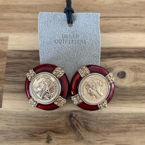 May include: A pair of red and gold-toned earrings with a coin-like design. The earrings have a circular shape with a gold coin in the center and are labeled with the text "Urban Outfitters".