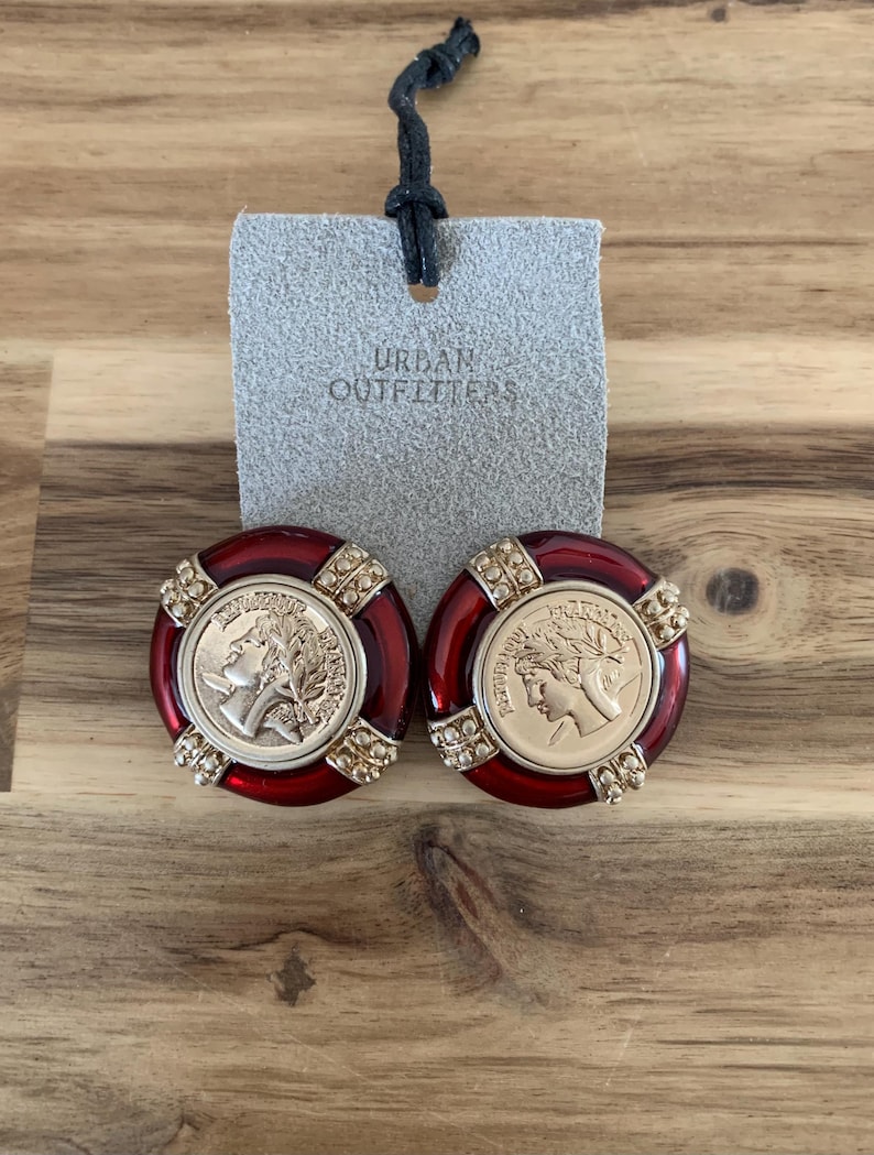 May include: A pair of red and gold-toned earrings with a coin-like design. The earrings have a circular shape with a gold coin in the center and are labeled with the text "Urban Outfitters".