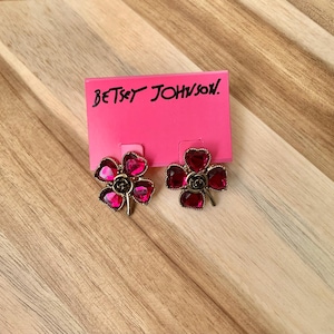 Könnte beinhalten: Ein Paar goldfarbene Ohrringe mit rosafarbenen Blumendesigns. Die Ohrringe haben die Form eines vierblättrigen Kleeblatts mit einer Rose in der Mitte. Die Ohrringe sind von der Marke "Betsey Johnson."