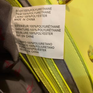 May include: A bright neon yellow handbag with a grey interior lining. A white care label is attached to the inside, displaying material composition in English, French, and Spanish, stating 100% polyurethane body and trim with 100% polyester lining.
